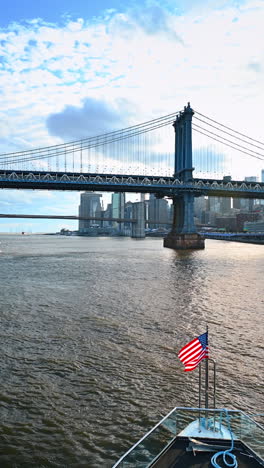 travelling on board of the boat with american flag at front by the east river. the manhattan bridge and the brooklyn bridge. vertical video
