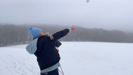 Toma-En-Cámara-Lenta-De-Una-Joven-Atractiva-Lanzando-Bolas-De-Nieve-Al-Aire-Durante-Un-Hermoso-Día-De-Invierno-Nevado-Con-Bosque-Y-Niebla-En-El-Fondo