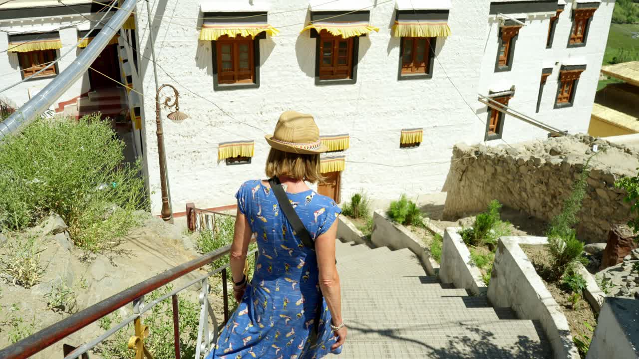 A blonde haired woman walks down stairs outside Monastery, Leh, in a serene, sunny setting. Exploring temple in blue dress on an adventure holiday visiting religious sites.