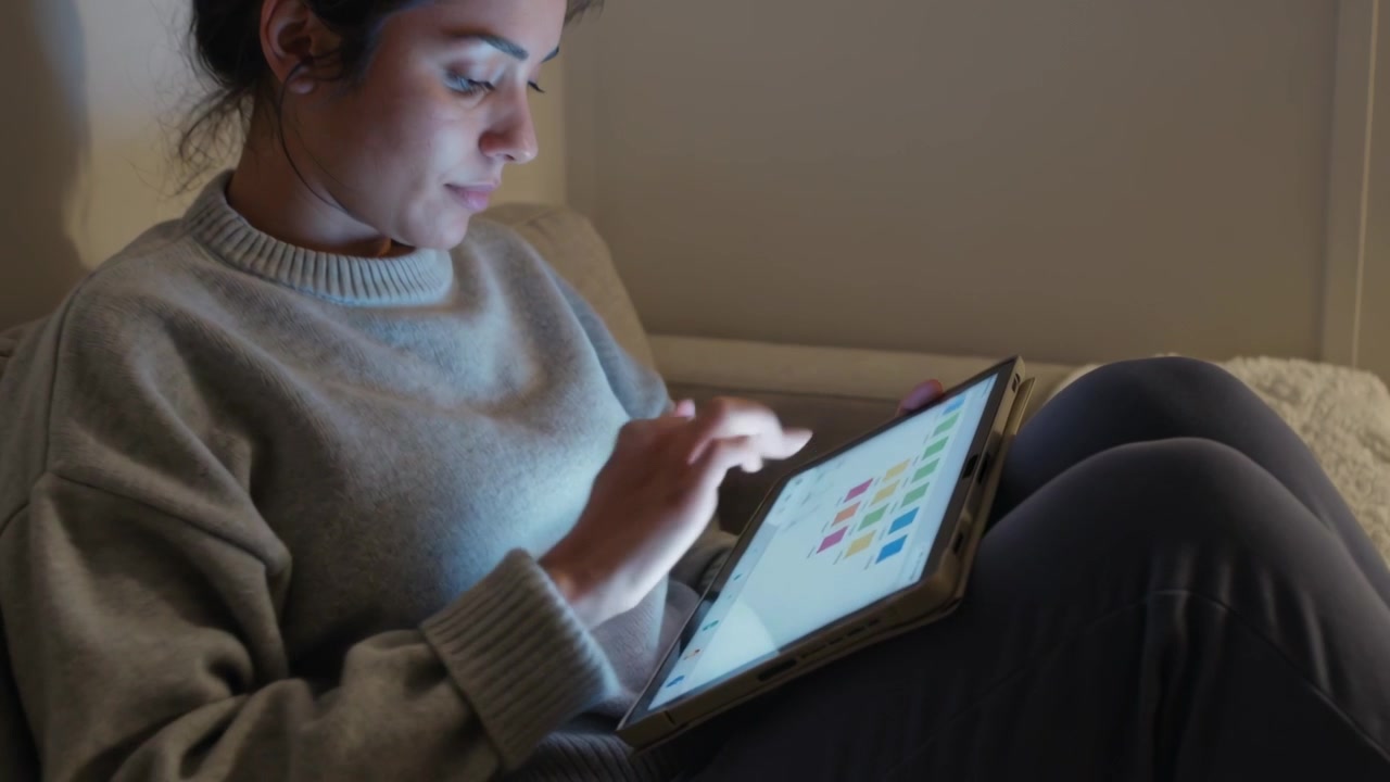 Tapping woman in knit sweater sitting on sofa at home, tablet lighting face selecting colored tiles