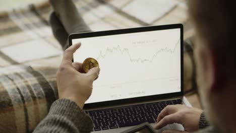 trader trading bitcoin, crypto currency chart on laptop screen and golden coin in hand, closeup view