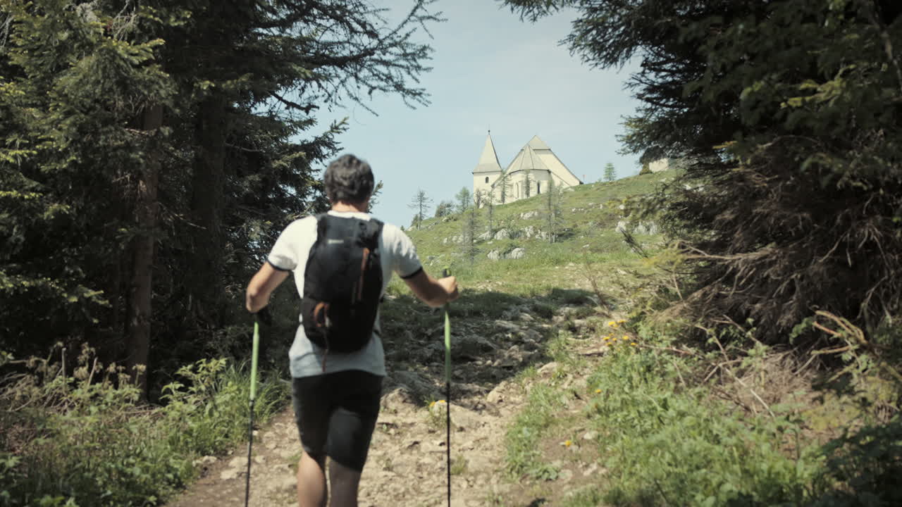 excursionista subiendo una colina, caminando con bastones de senderismo pasando las coníferas y hacia la iglesia que se encuentra en la cima de la montaña