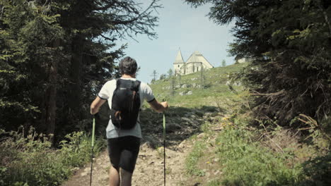 hiker climbing up a hill, walking with hiking poles past the conifers and towards the church which is standing at the top of mountain