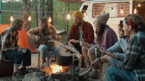 young woman playing guitar while sitting with friends by campfire