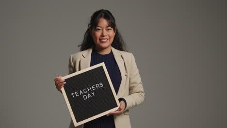 studio portrait of female teacher standing against grey background holding notice board reading teachers day