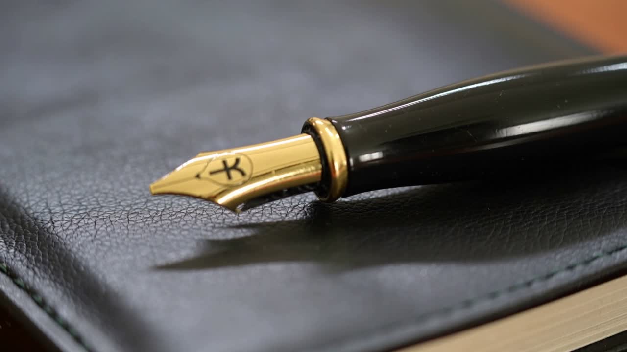 Ajustando o foco, close-up de caneta-tinteiro com pena de ouro repousando sobre caderno de couro preto, mostrando uma marca.