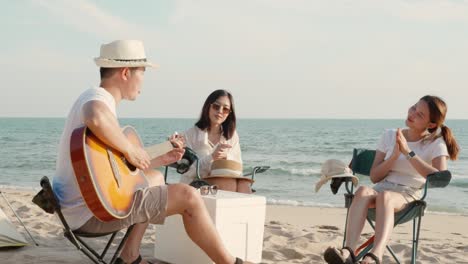행복한 친구는 캠핑 텐트 근처의 모래 해변에서 휴가를 함께 미소 짓고 캠핑에서 기타를 연주하고 박수를 치며 즐거움을 얻습니다.