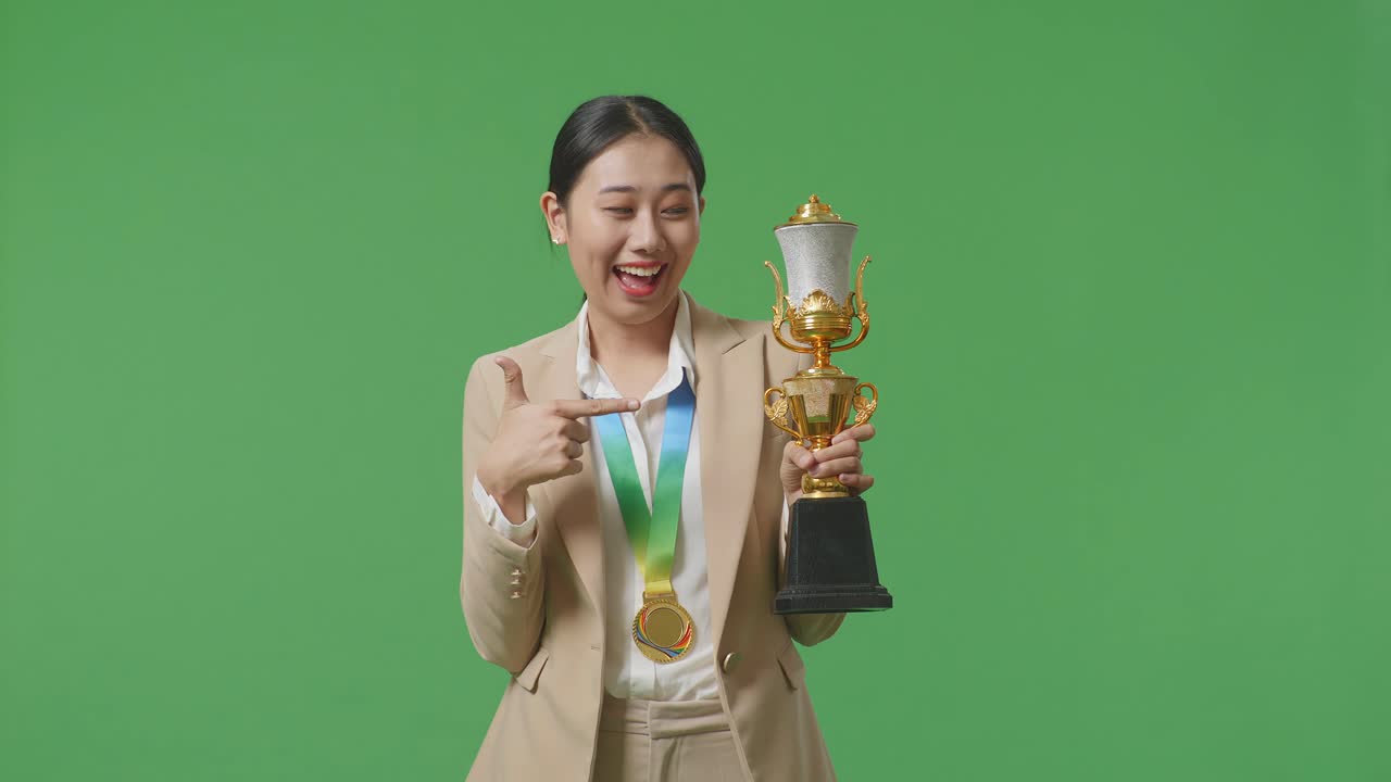 mujer de negocios asiática en traje con una medalla de oro señalando un trofeo de oro en su mano estando orgullosa de ganar como la primera ganadora en el fondo de la pantalla verde en el estudio