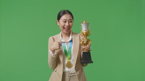 금메달을  슈트를 입은 아시아 사업가 여성은 손에 금 트로피를 가리키며 스튜디오의 녹색 스크린 배경에서 첫 번째 우승자로 자랑스럽게 승리합니다.