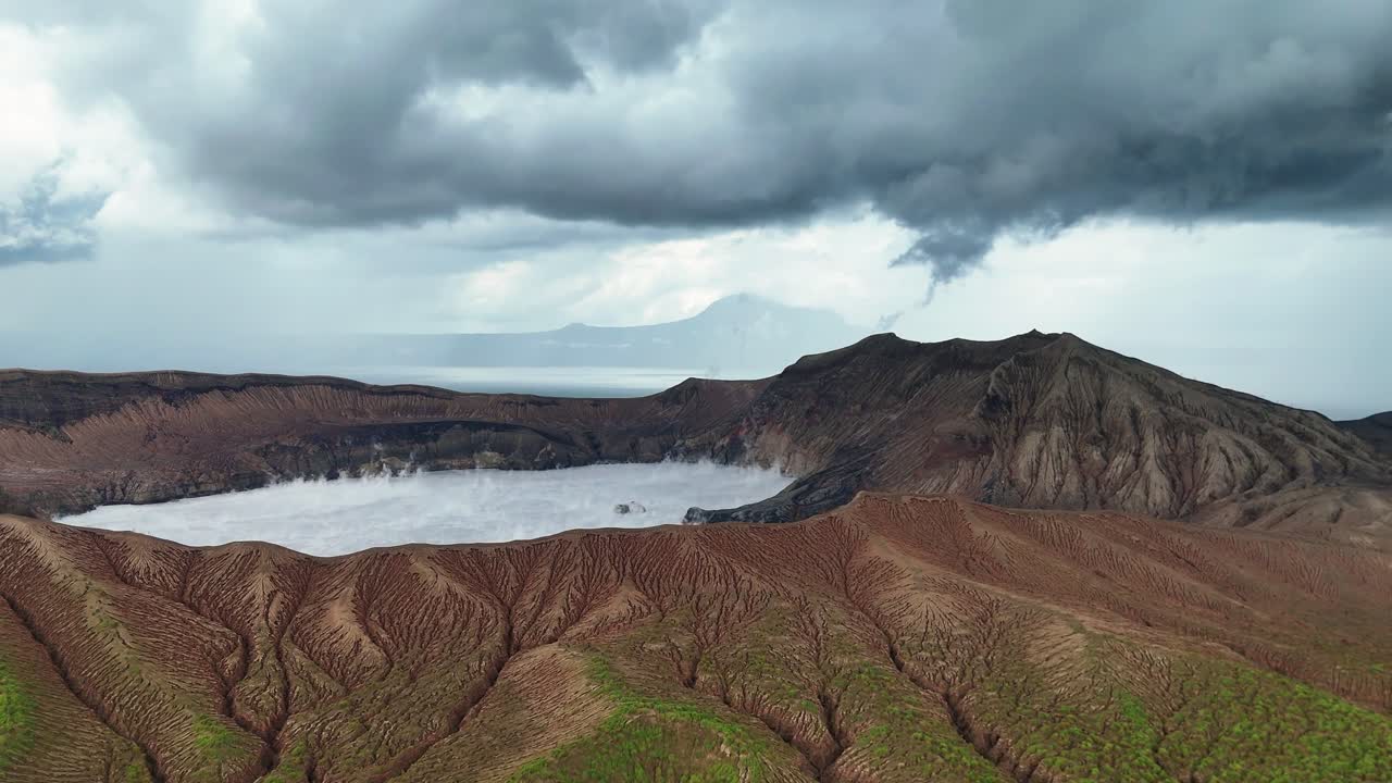 Wide aerial shot over the crater of Taal Volcano in the Philippines, with steam visibly rising from the opening, surrounded by dark, stormy clouds that add a dramatic effect.