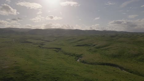 slow drone push-in capturing a sunset over grassy fields ascending a mountain, with a tiny stream carving a path from melting snow