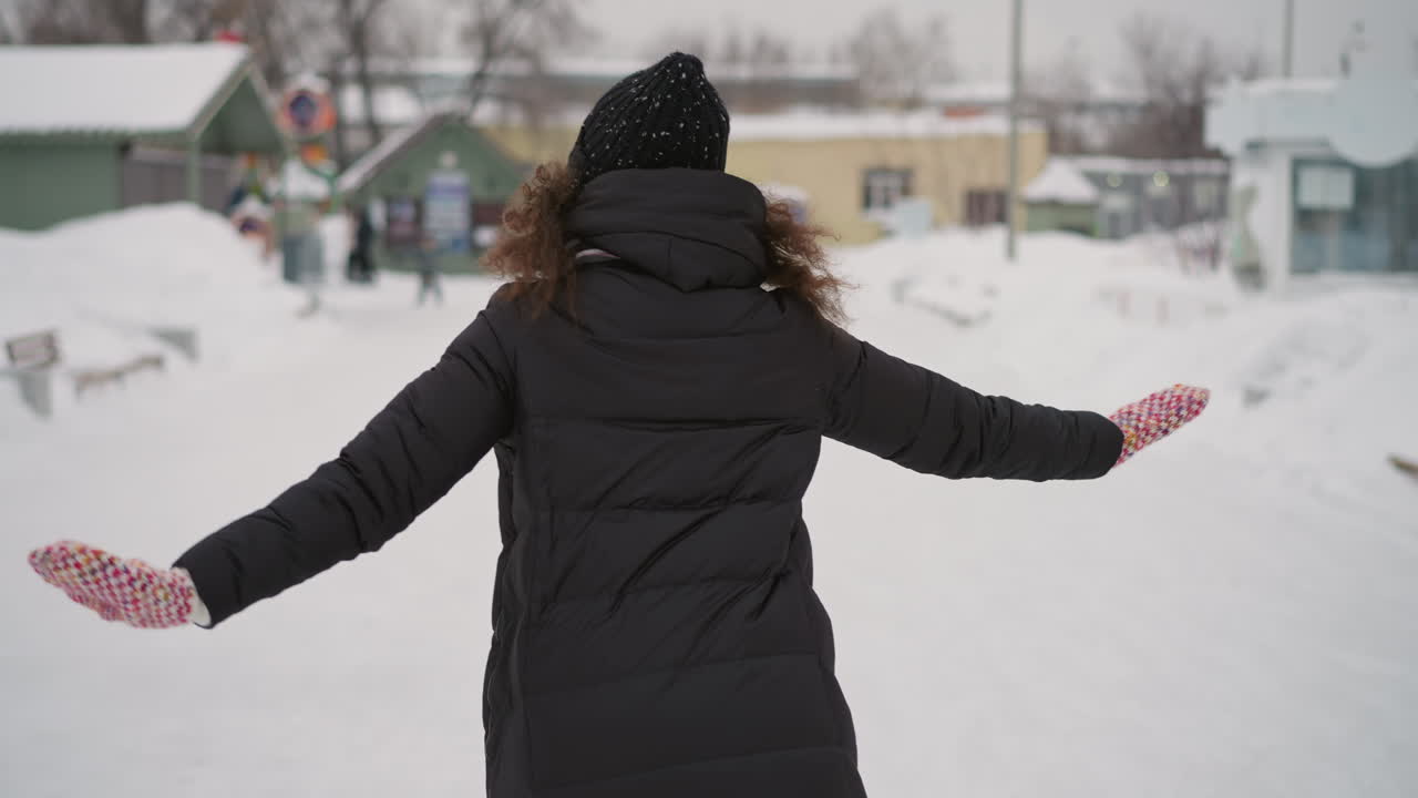 Athletic girl dressed in long winter coat, knit hat, and colorful mittens outdoors with arms spread wide on snowy street, enjoying cold season atmosphere with playful carefree mood in frosty environment