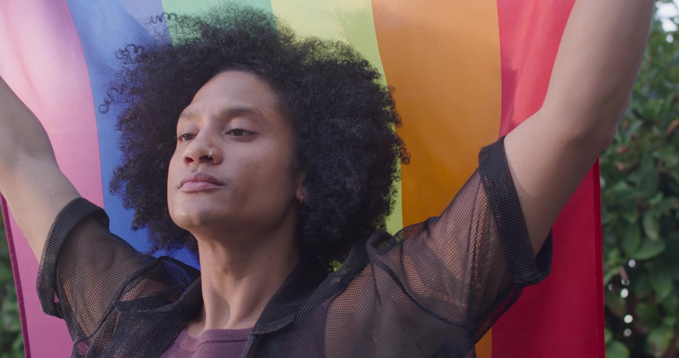 Uomo afroamericano adulto che inizia tenendo una bandiera arcobaleno, alzando le braccia, mostrando orgoglio in giardino.