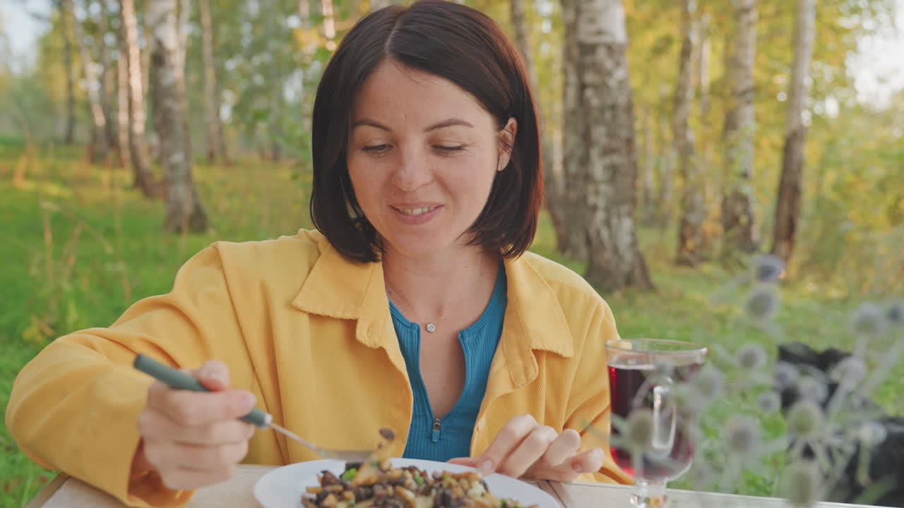 Mujer caucásica acampando en una mesa de forrajeo disfrutando de un desayuno en el bosque entre abedules, vajilla rústica con ensalada y sartén, copa de vino cerca, expresión pacífica saboreando un bocado, prado soleado.