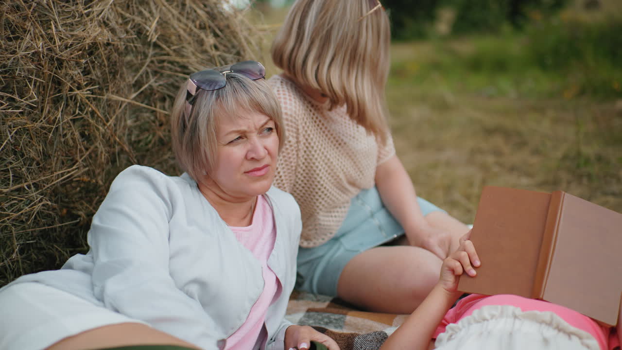 가족은 야외에서 조용한 순간을 즐기고, 야외에서 풀 근처에서 휴식을 취하고, 사려 깊은 여자가 멀리 쳐다보고, 다른 한 명은 뒤로 기대고, 아이는 읽기 위해 누워 있습니다.