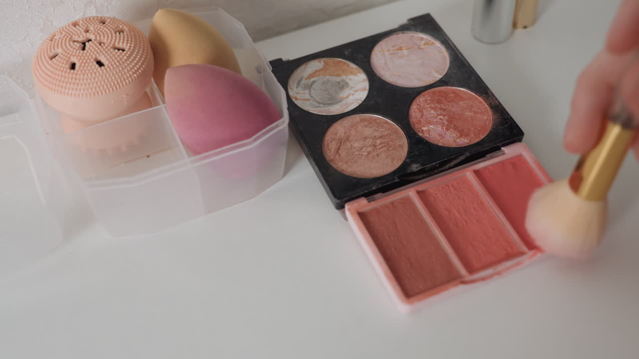 Pink makeup palette brush sponge closeup, hand sweeping blush pan in soft glam demo, pastel shimmer eyeshadow and peach blush pans, translucent container and sponge, textured powder particles, cozy