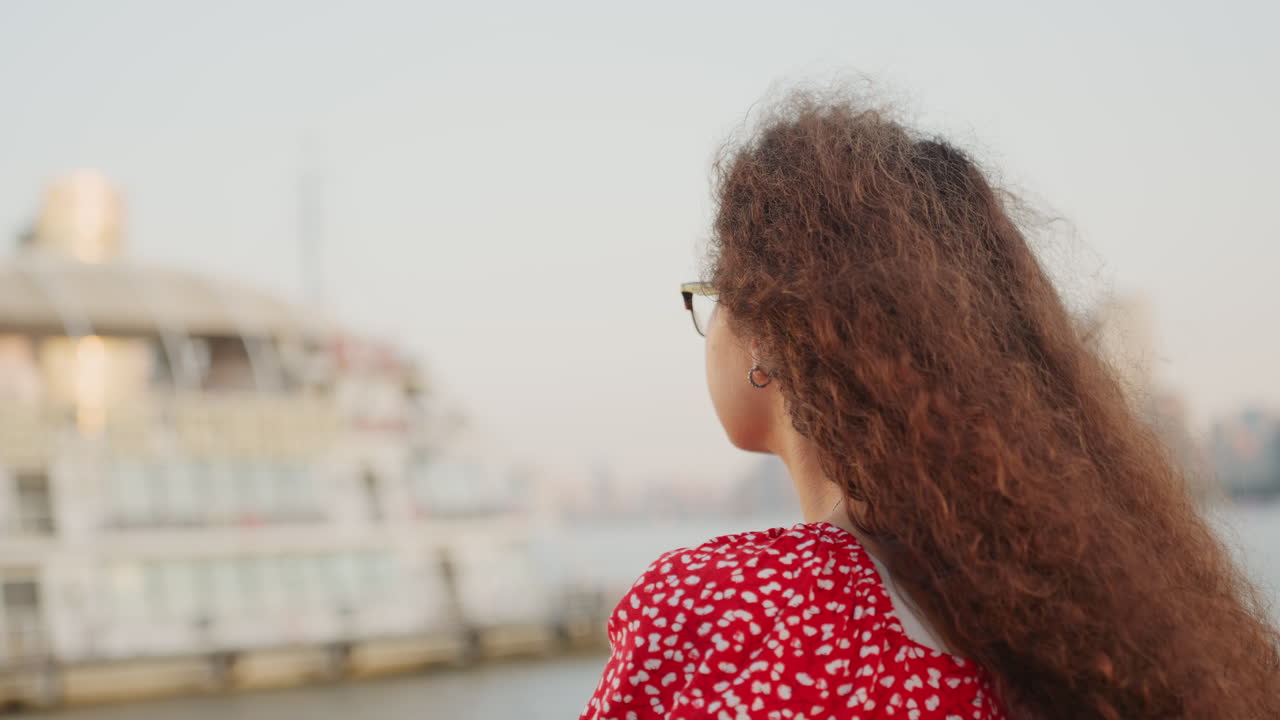 Woman looking at ferry waterfront skyline, backview in red floral dress with glasses, long curly hair blowing in breeze, soft evening light on dock and moored boat, calm urban harbor atmosphere