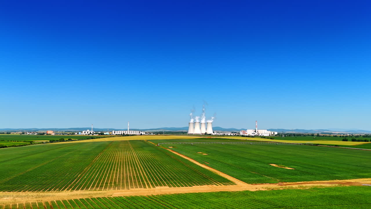 Flight over green plantations in the countryside approaching the irrigation system. Huge white pipes of the plant produce smoke
