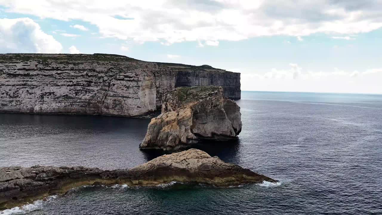 Fungus Rock Il-Ġebla tal-Ġeneral stands in Dwejra Bay San Lawrenz Gozo Malta beneath towering Lower Coralline Limestone cliffs along Mediterranean coast, aerial drone push forward shot