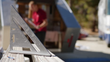 A-wood-worker-placing-a-carpenters-square-on-a-bench-as-a-construction-worker-builds-a-travel-trailer-in-the-background-with-copy-space