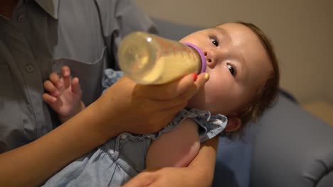 Hungriges-Kleines-Mädchen-Auf-Den-Armen-Der-Mutter,-Das-Milch-Aus-Plastikflasche-Trinkt,-Nahaufnahme