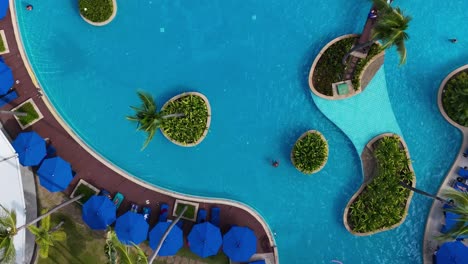 drone captures a vibrant pool with unique palm-shaped islands and blue umbrellas, creating a tropical oasis.