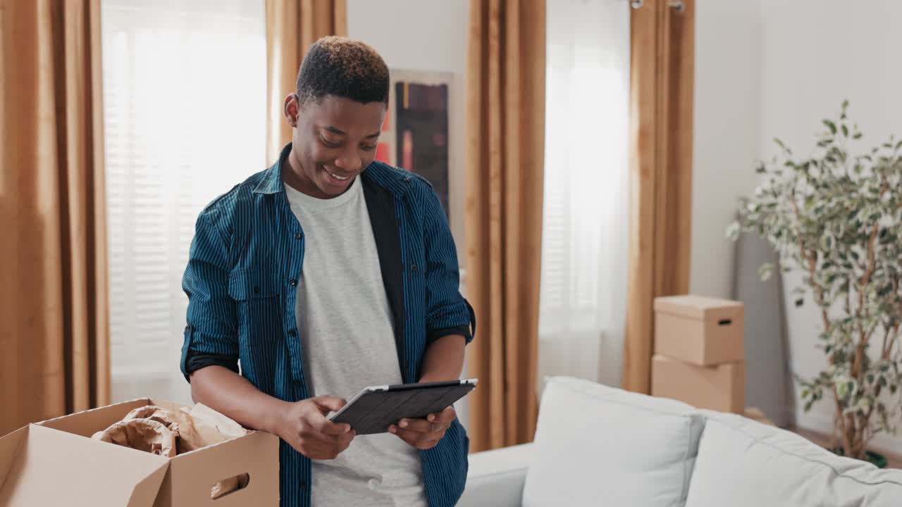 A young boy hangs out in his new apartment, unpacking cardboard boxes left over from the move, browsing the web on tablet, social media, laughing at memes, browsing inspiration