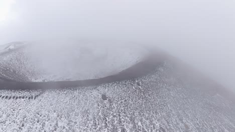 an aerial shot of one of mount etna's extinct craters, with the cone of the crater clearly visible. dense fog surrounds the area.