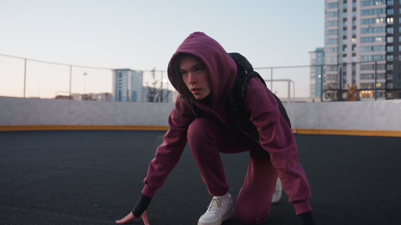 Athlete kneeling in starting sprint position on black asphalt sports court next to white barrier topped with chain link fence in urban environment wearing maroon hoodie black vest and white sneakers