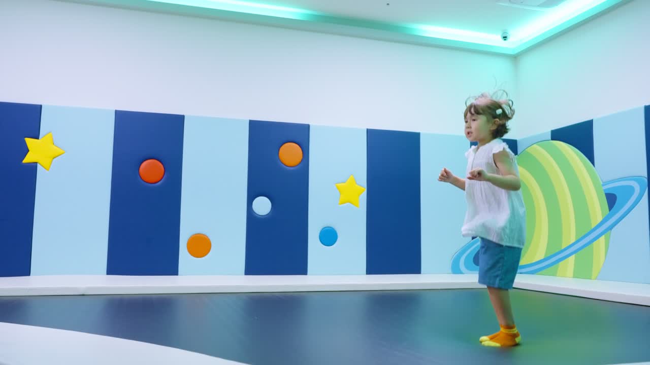 Gadis kecil yang bahagia bersenang-senang melompat di trampolin di taman bermain dalam ruangan penuh warna bertema luar angkasa.