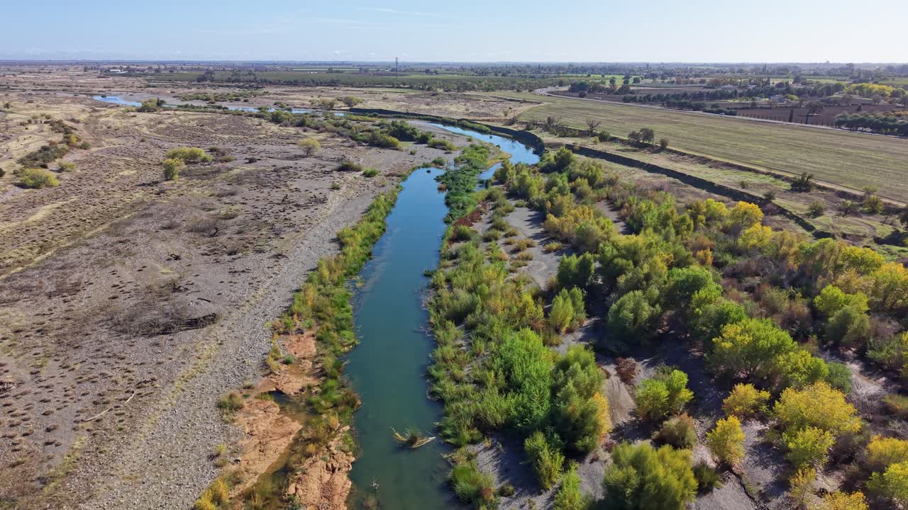 Drone flies forward at constant height above Cache Creek, showing dry riverbanks, green trees and farmland in Capay, California