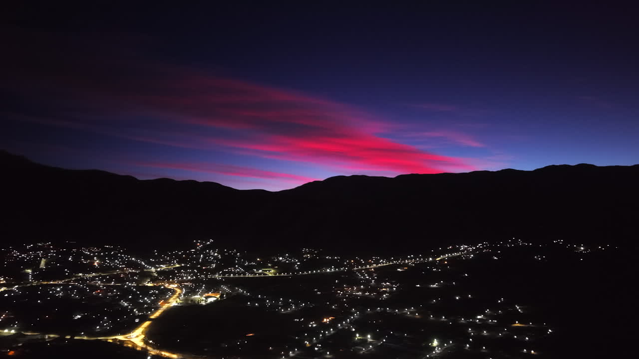Sunrise soft glow above the silhouetted mountains and sparkling town lights in Tafí del Valle, Tucumán Province, Argentina, aerial drone slow pan