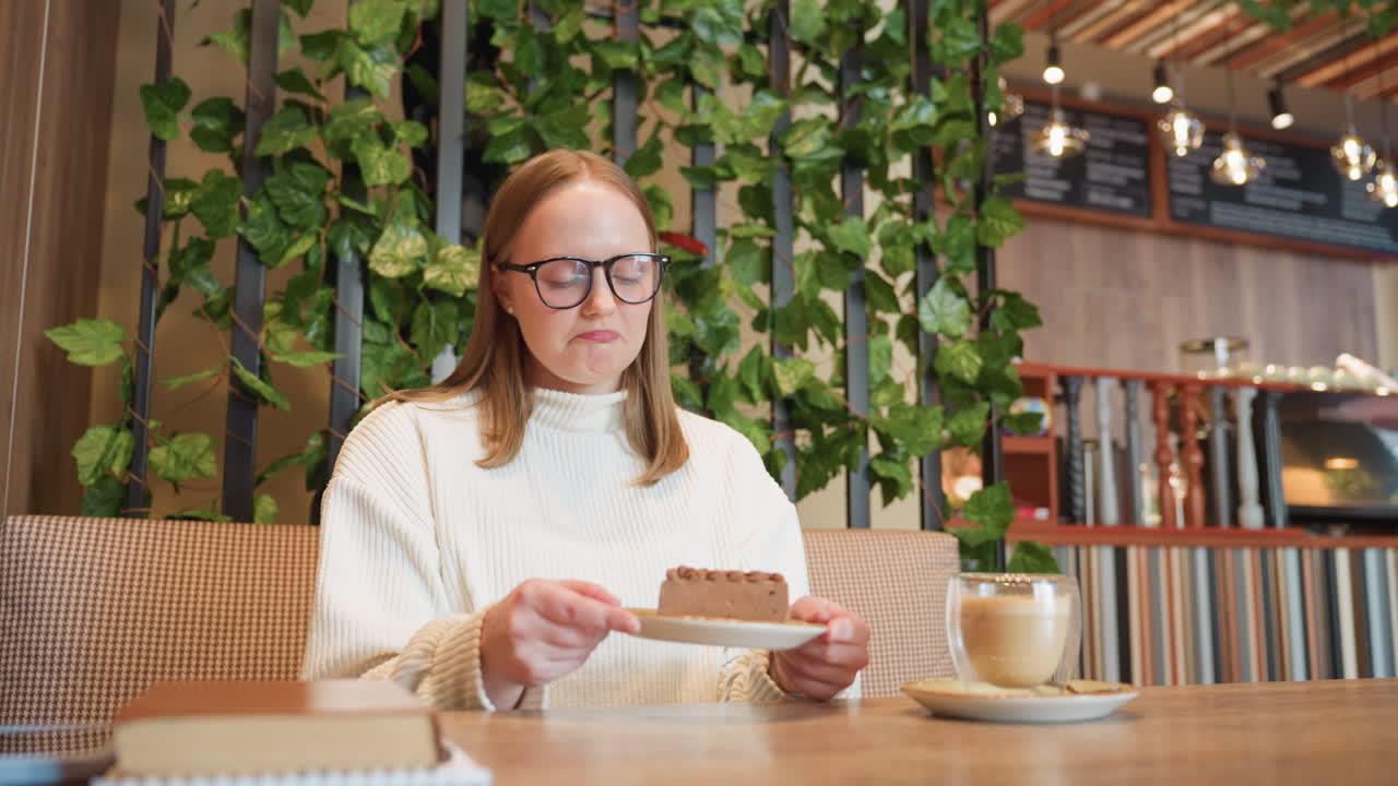 Lady seated at cozy cafe table holds chocolate cake close to nose, smiling as she smells dessert, with latte on table and green leafy decor in background