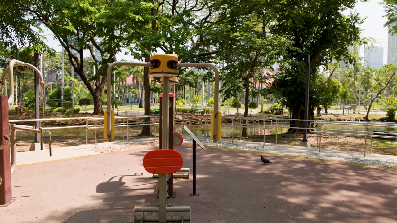 Toma panorámica estática de máquinas de ejercicio vacías bajo árboles verdes en un parque público soleado