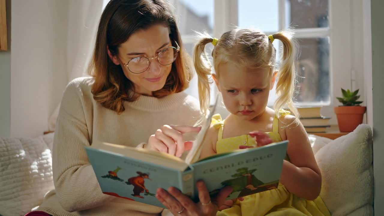 Madre che apre un libro e guida la figlia in vestito giallo, girando le pagine e indicando per imparare sul divano.