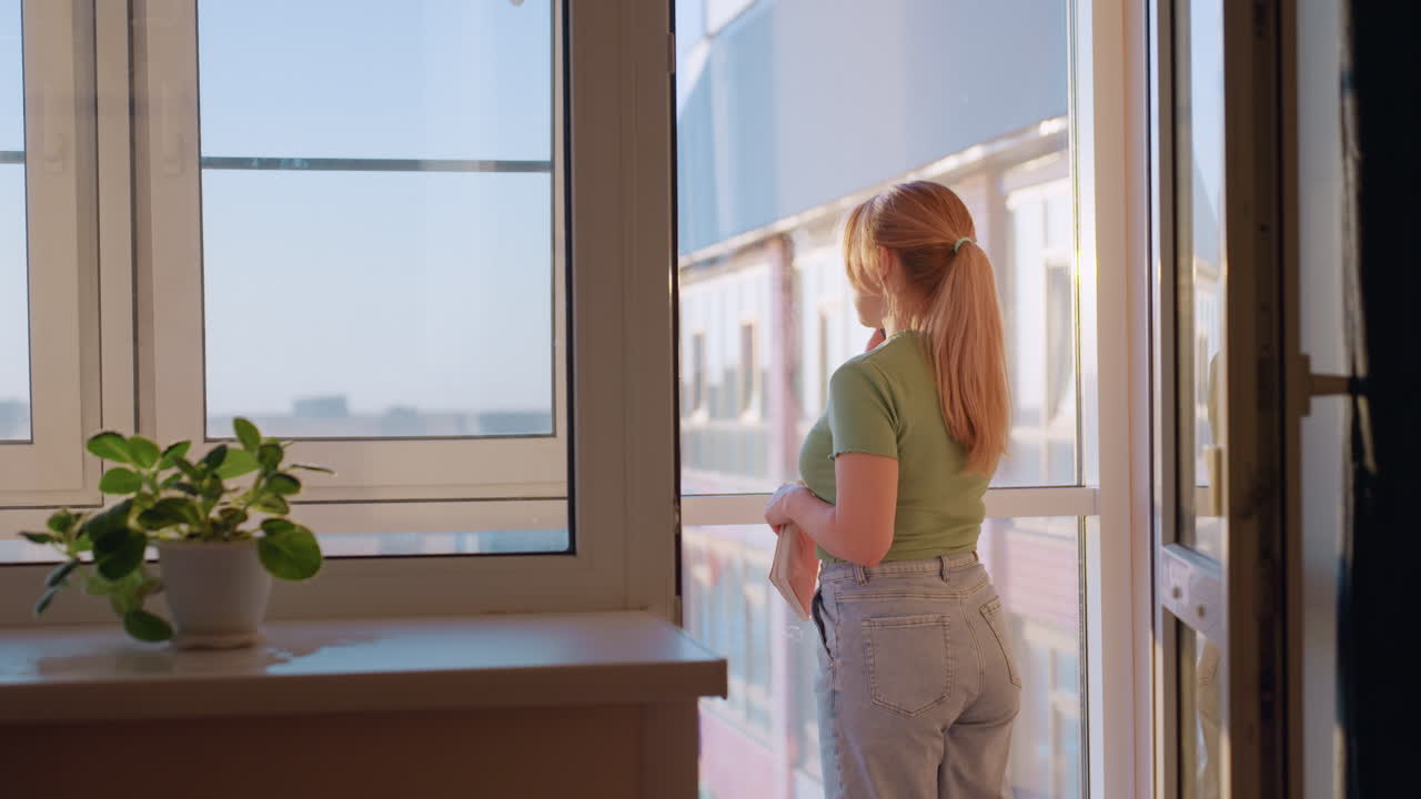Lady with ponytail wearing green shirt walks toward balcony while on call, standing near bright window with potted green plant on windowsill, sunlight casting warm glow across home