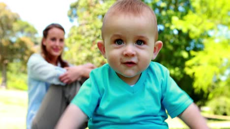 un bebé riendo sentado en el césped con su madre detrás de él