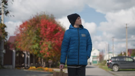 young boy in blue jacket and black beanie strolls thoughtfully looking around in residential area, sunlight reflecting on his face, with autumn trees and parked car in the background