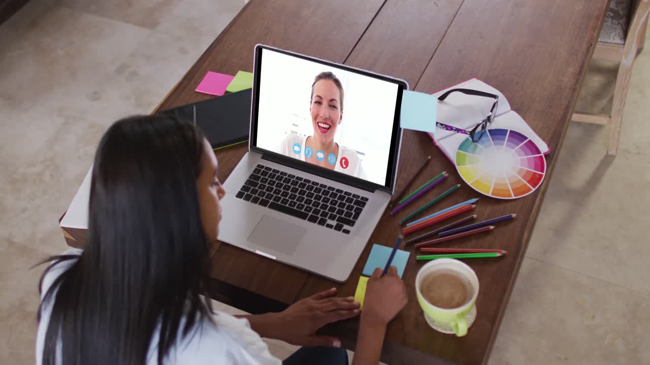 Mujer caucásica usando una laptop en videollamada con una colega, tomando notas