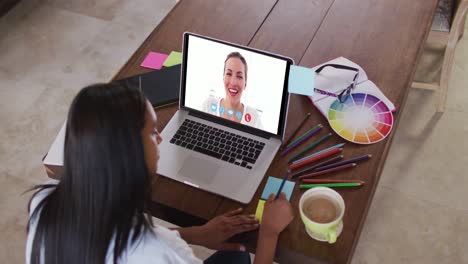 mujer caucásica usando una computadora portátil en una videollamada con una colega, tomando notas