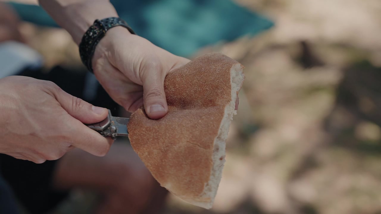 Hands cutting a crusty bread roll open with a pocket knife at an outdoor gathering