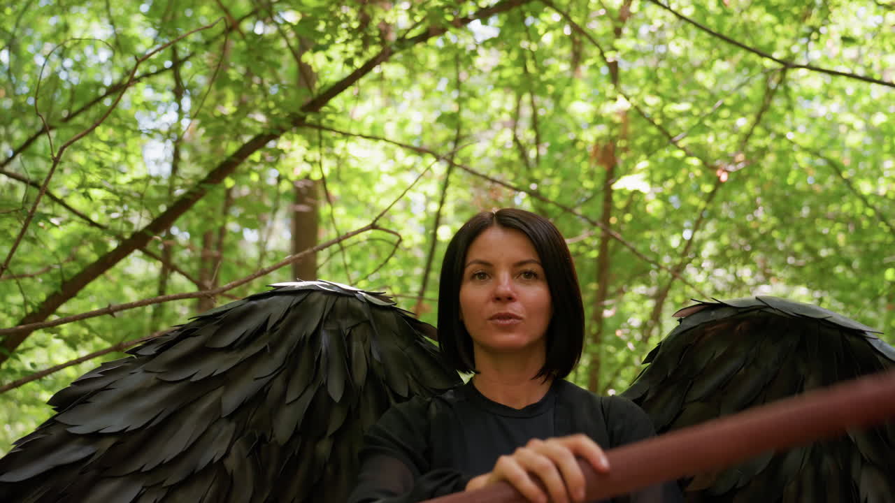Young lady dressed in dark wings rotates staff gracefully in sunlit forest, surrounded by lush green leaves and glowing natural light, capturing mystical motion