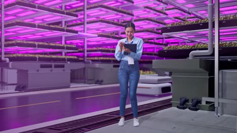 cuerpo lleno de una trabajadora profesional asiática de pie con su tableta en una granja vertical, instalación industrial de alta tecnología, escribiendo en el teclado de su tableta con meditación