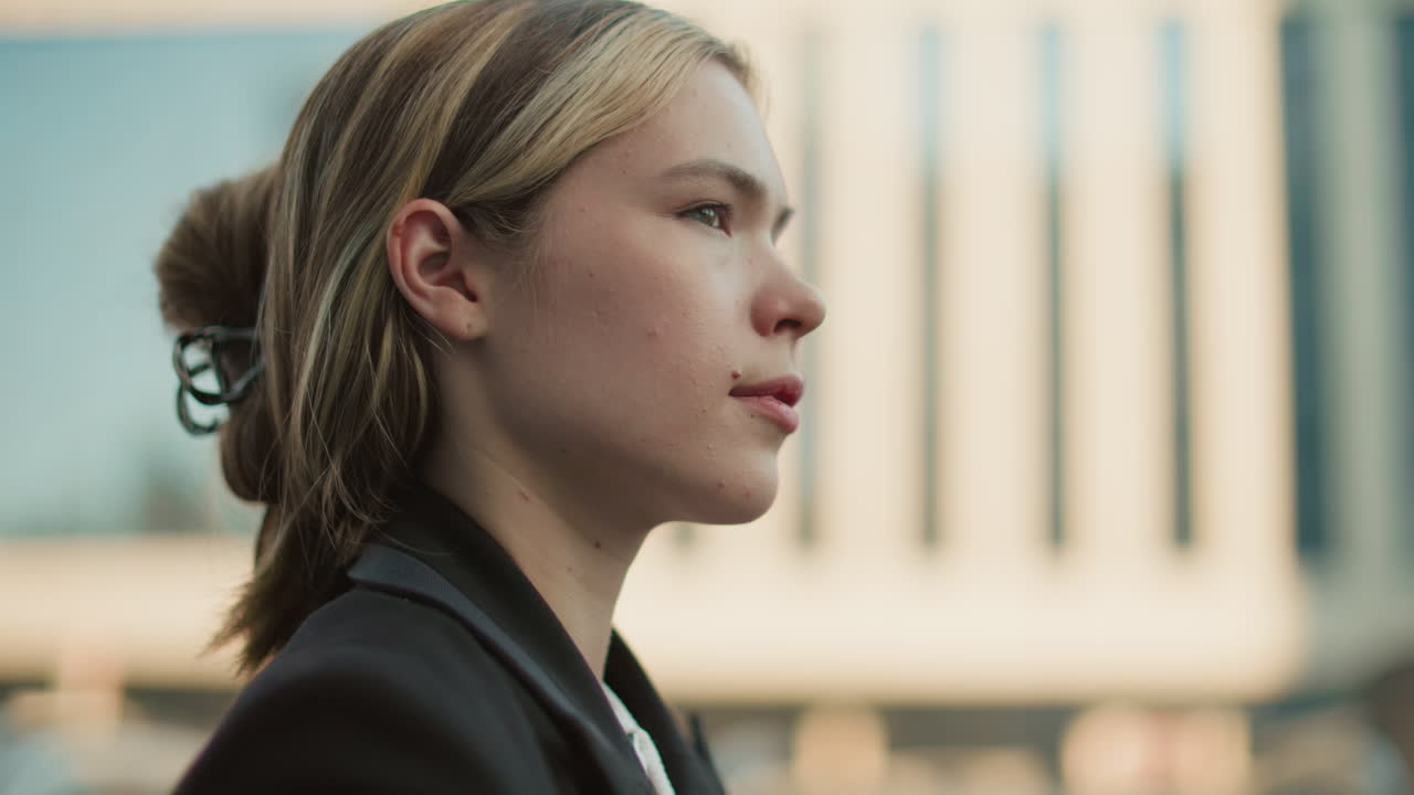 Close up side view of female stylist in black suit walking with confident expression and hair clipped back, blurred background of city buildings in urban setting during daylight workday moment