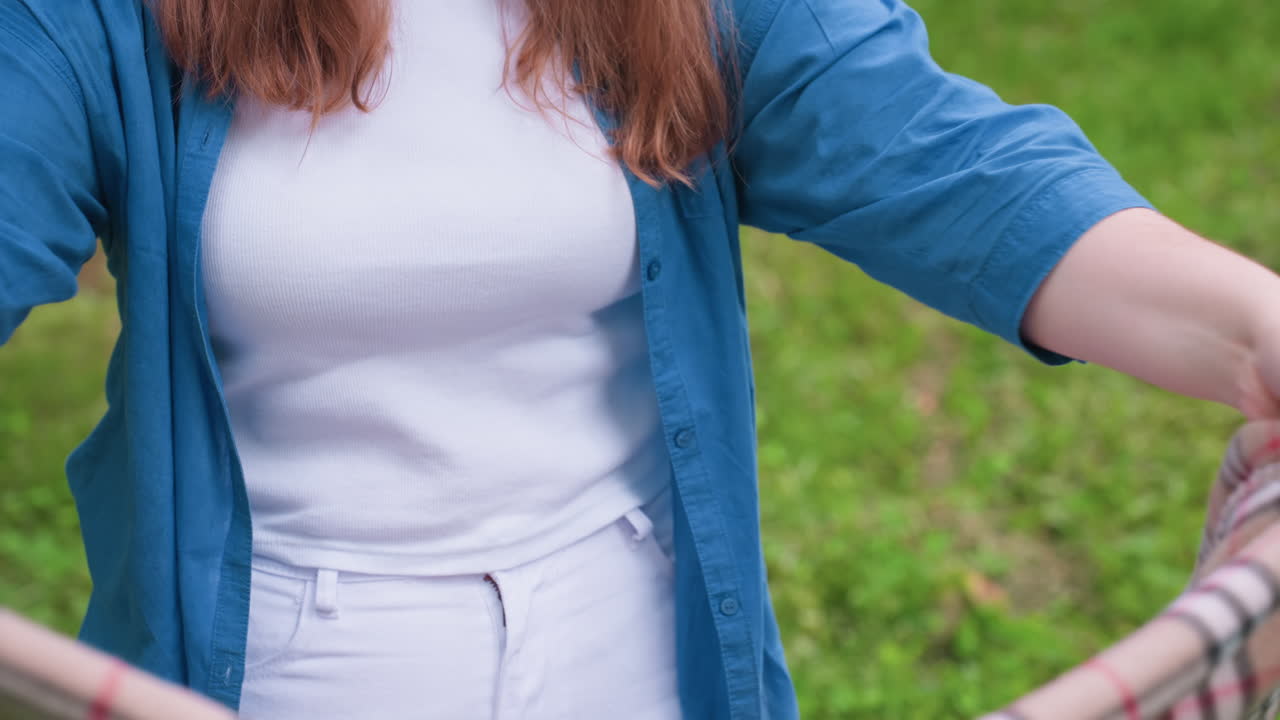 Close up of student folding blanket carefully while standing on green grass, dressed in casual blue shirt and white top, enjoying calm outdoor moment surrounded by natural greenery