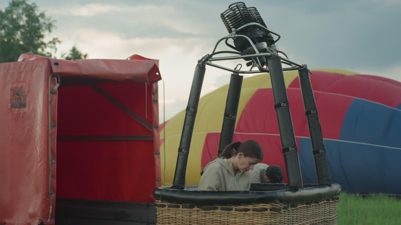 woman in light jacket stands inside wicker basket inspecting burner assembly while deflated red blue balloon envelope lies on grassy field under cloudy sky as team prepares for sunrise flight launch