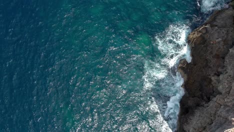 Top-Down-View-Over-A-Rocky-Shore-Line-In-Malta,-With-Waves-Splashing-And-Turquoise-Crystal-Clear-Water