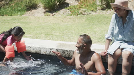 una chica biracial se inclina sobre el borde de una piscina hacia un hombre biracial en el agua