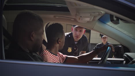 serious police officer checking black men in car zoom in view of serious man in police uniform asking questions and checking documents of african american men in modern vehicle on city street
