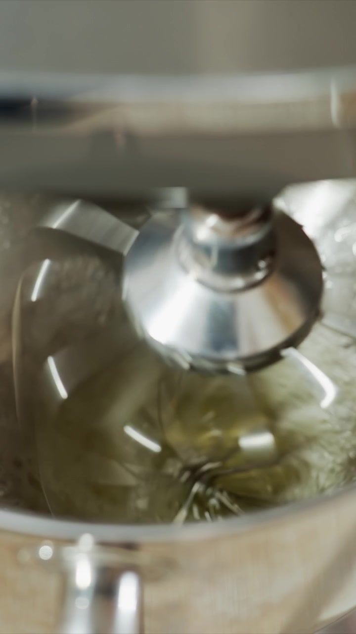 Close up of stand mixer whisk blending green liquid. Mixture thickens and froths as it is continuously mixed. Kitchen countertop edge appears briefly at frame bottom emphasizing